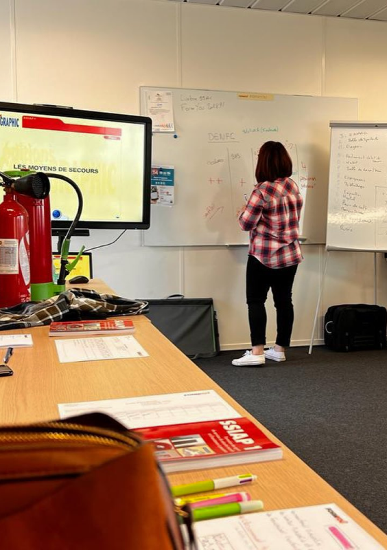 salle de formation avec un élève au tableau et des tables sur lesquelles sont disposés des supports pédagogiques